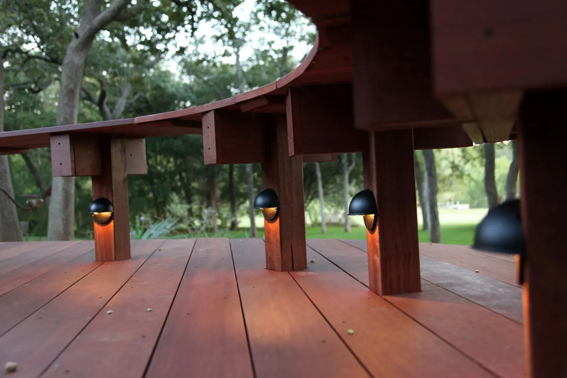 Wooden deck with warm lights illuminating the underside, set in a natural, green outdoor area.