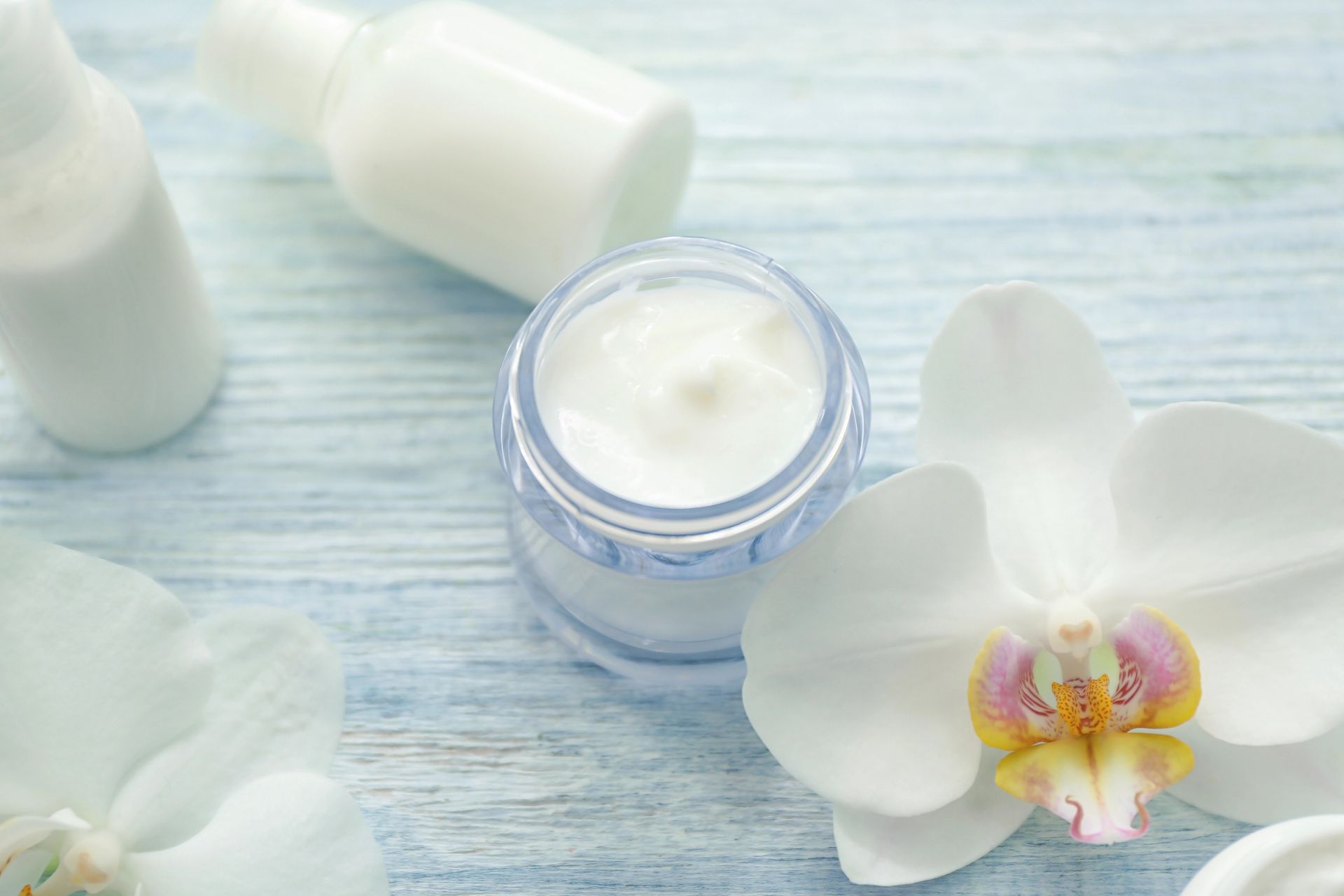 Barattolo di crema con lozione bianca, piccole bottiglie e orchidee bianche su una superficie di legno azzurro.