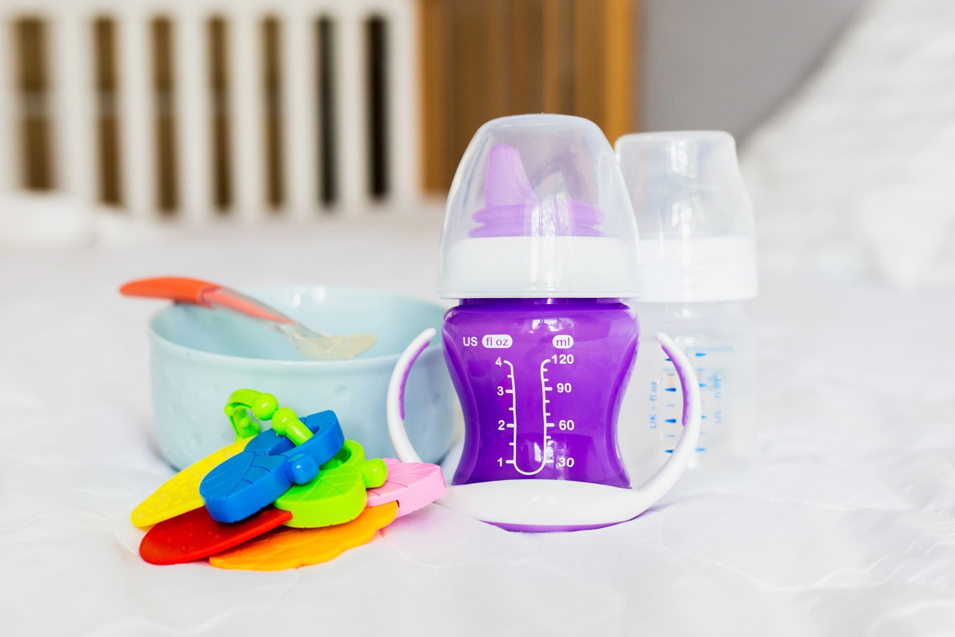 Biberon viola con manici, ciotola per il cibo e massaggiagengive colorati su un letto.