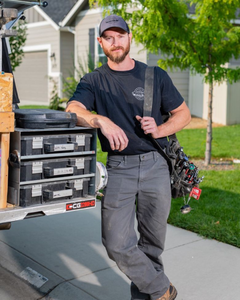 Man in work clothes leans on a van with tools, holding a bag. Residential setting.