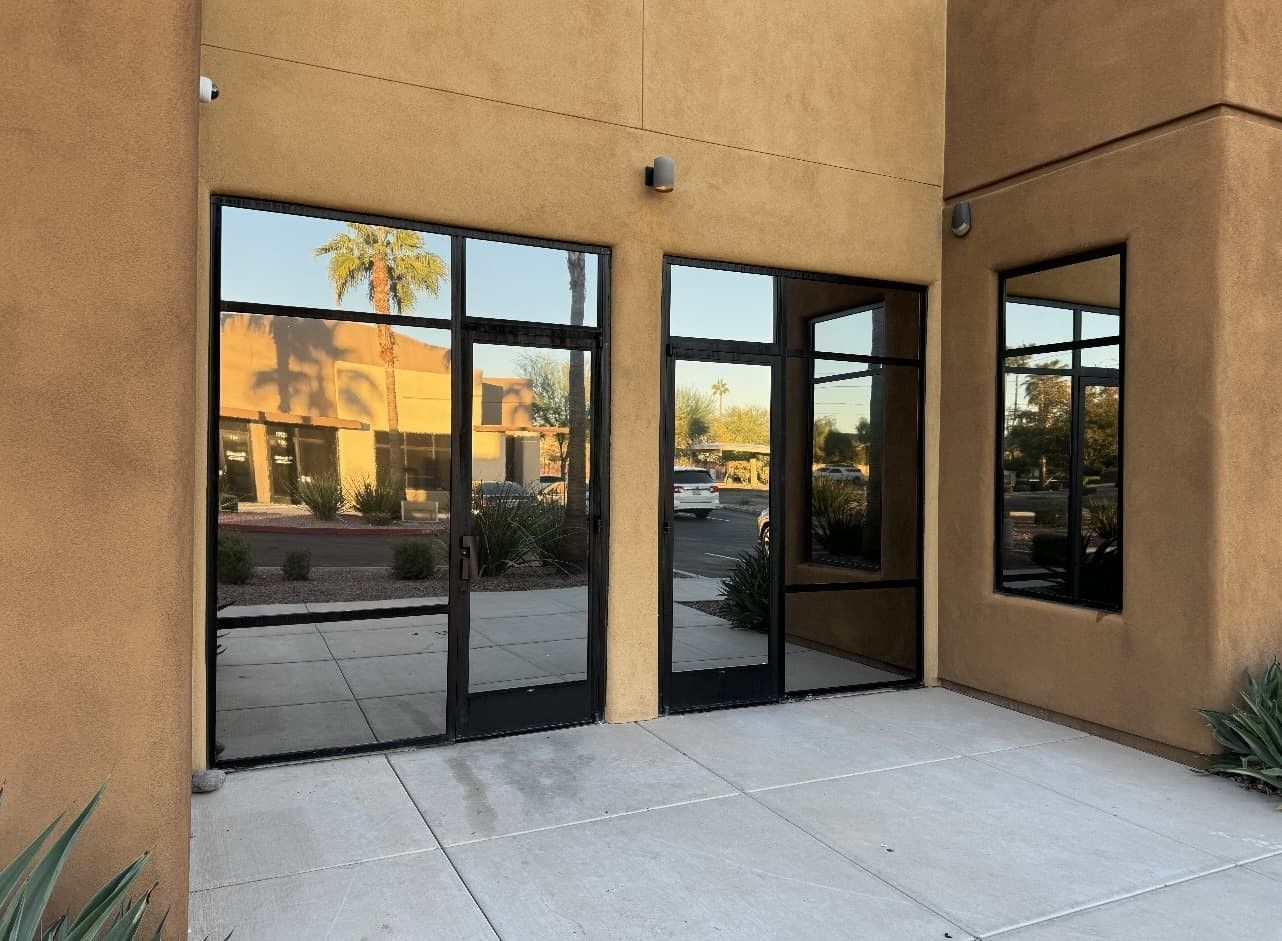 Modern glass storefront entrances in a tan stucco building with a concrete walkway and reflections of nearby trees.