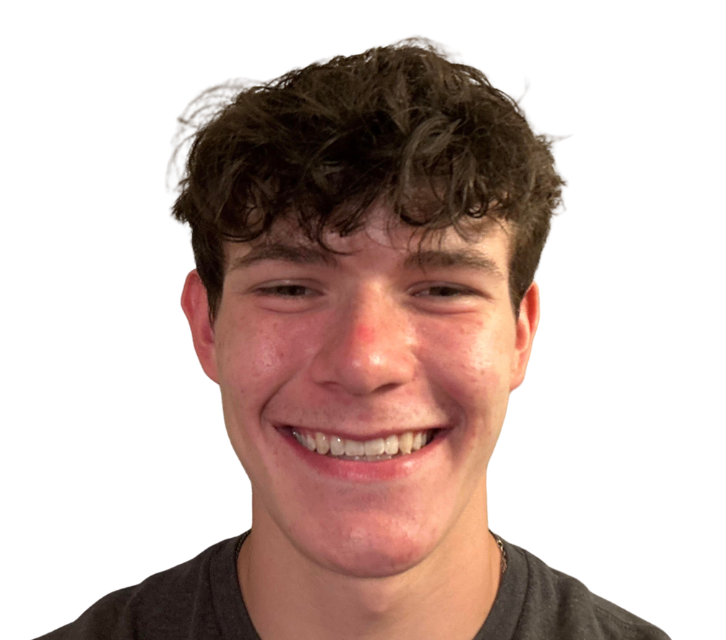 A young man with curly brown hair smiles widely. He wears a gray shirt and his face is flushed.