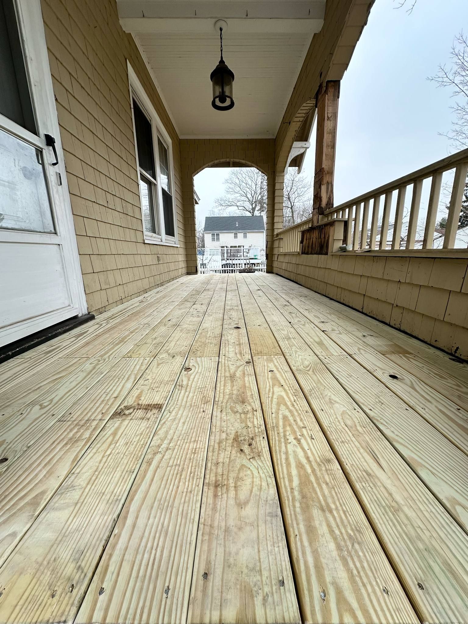Wooden porch with new planks, leading towards a house with railings on one side, overcast sky.
