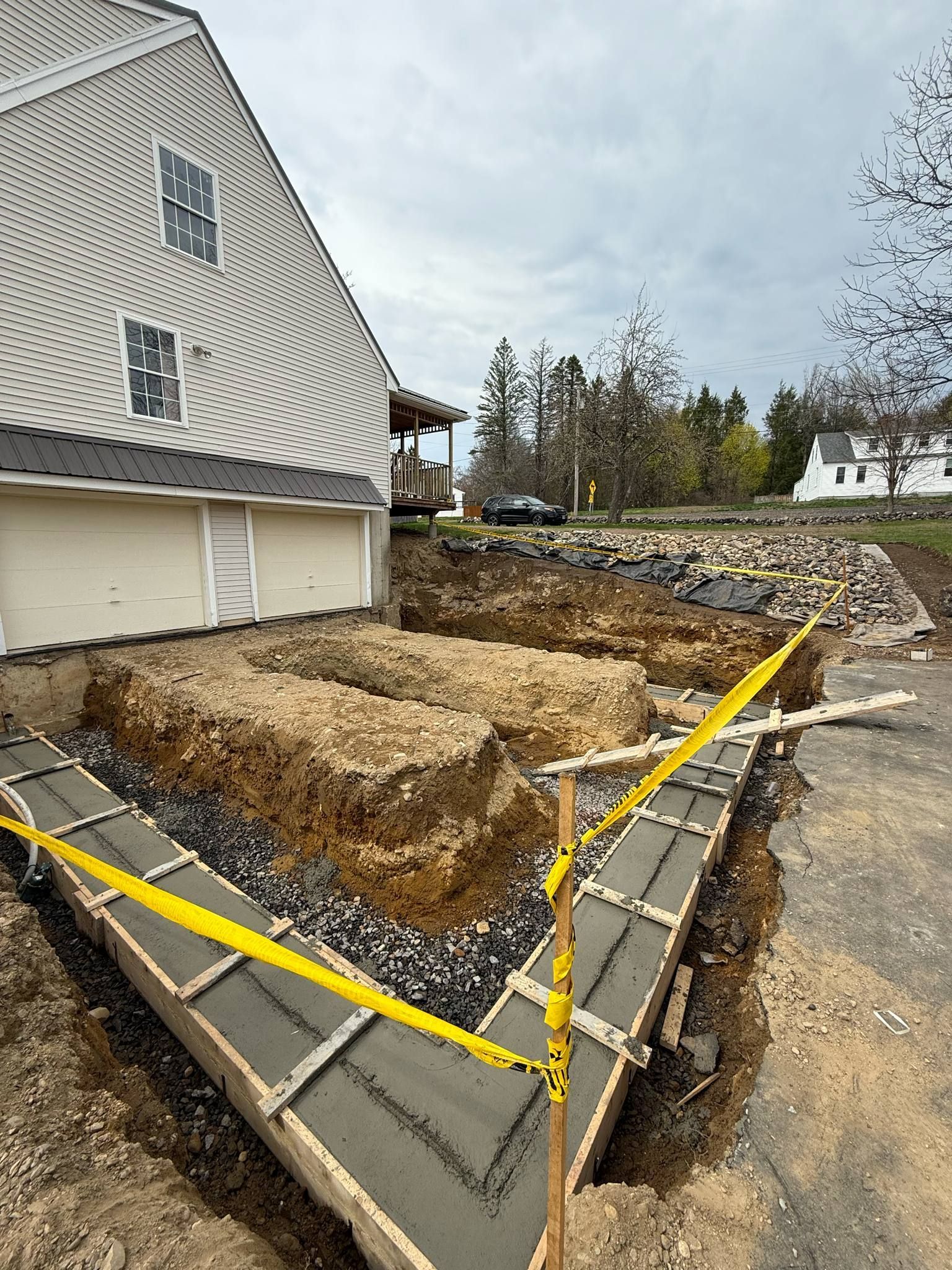 Construction site next to a house: freshly poured concrete forms, excavated soil, safety tape, and a cloudy sky.