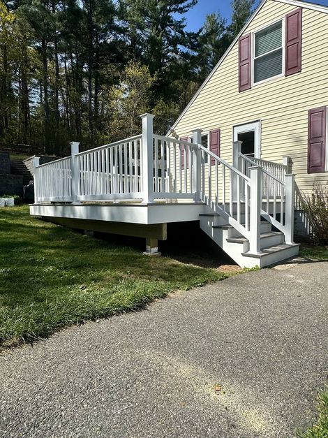 White deck with stairs attached to a yellow house, set in a grassy yard and gravel driveway.