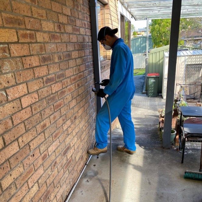 A Man in a Blue Jumpsuit is Spraying a Brick Wall — Country to Coast Pest Control In Urunga, NSW