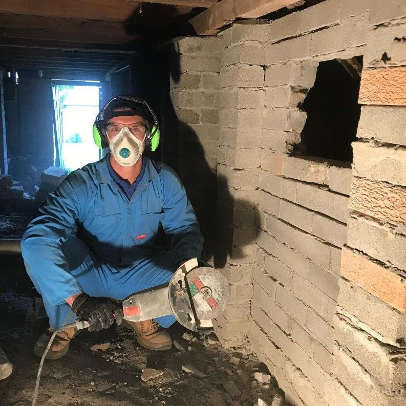 A Man Wearing a Mask is Kneeling Down in Front of a Brick Wall — Country to Coast Pest Control In Urunga, NSW