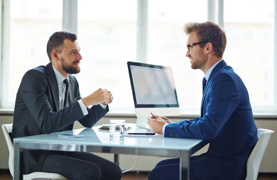 Zwei Männer in Anzügen sitzen an einem Tisch; einer spricht, der andere hört aufmerksam zu.