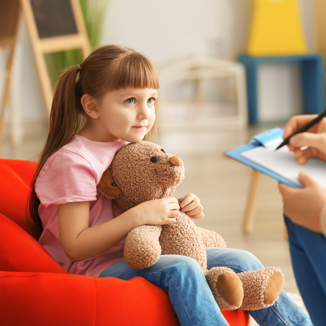 Young girl with pigtails hugs teddy bear, sitting on red beanbag chair; therapist taking notes.
