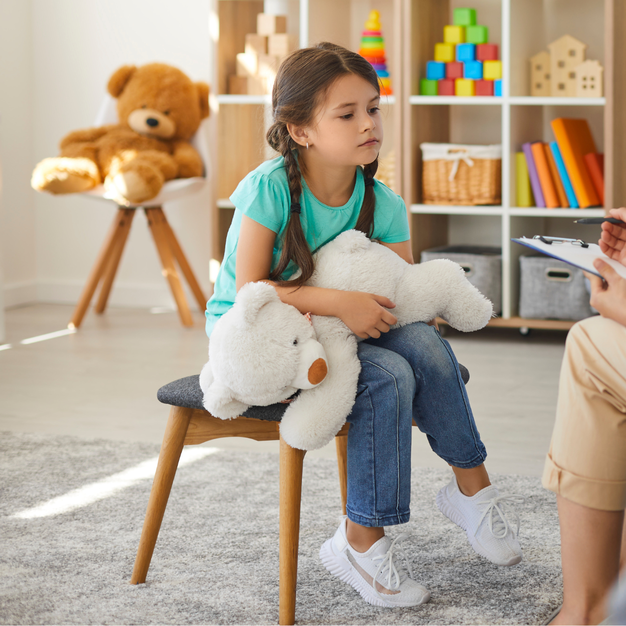 Girl holding a teddy bear in a therapist's office; listening to someone off-screen.
