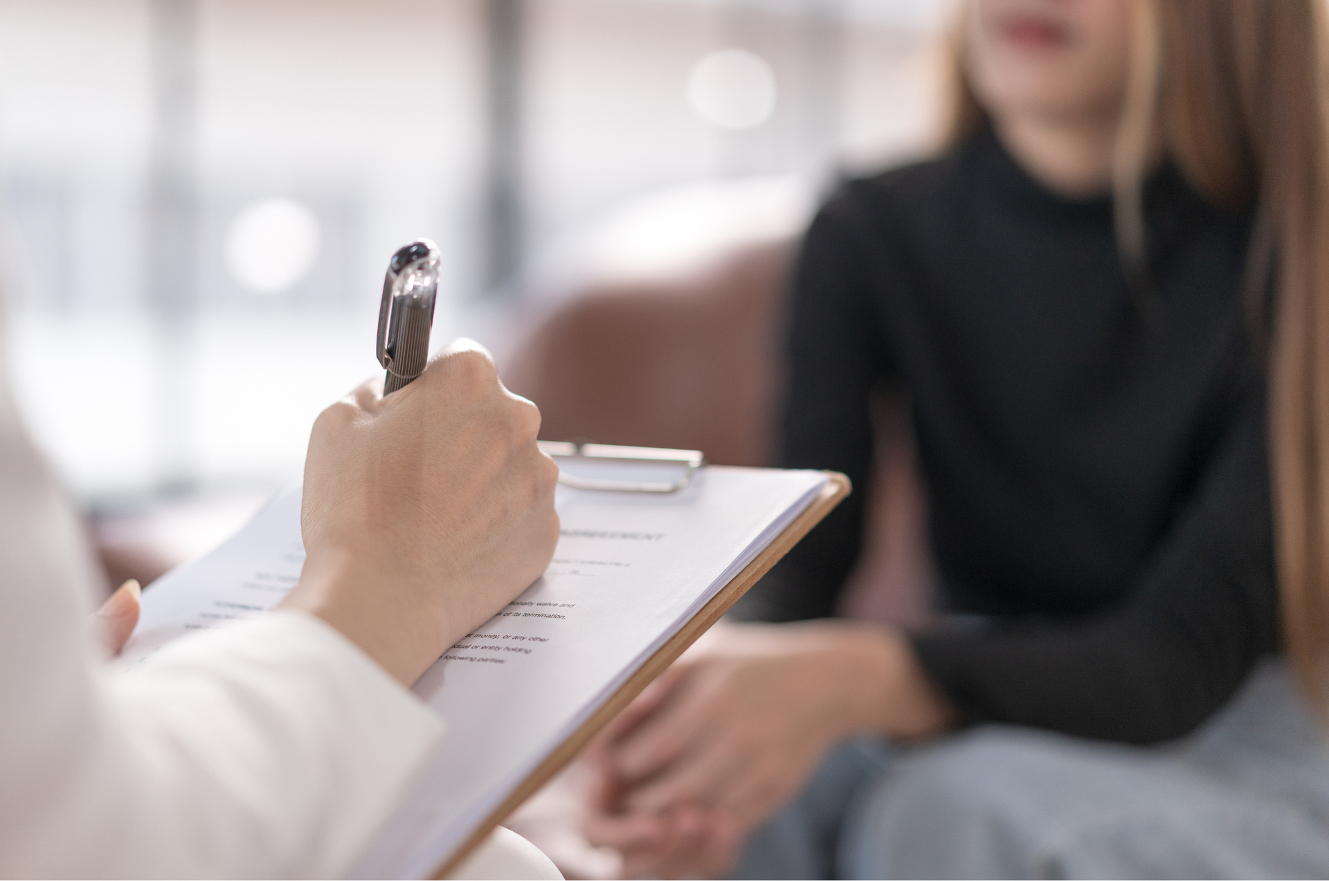 Therapist taking notes on a clipboard while speaking to a seated patient.