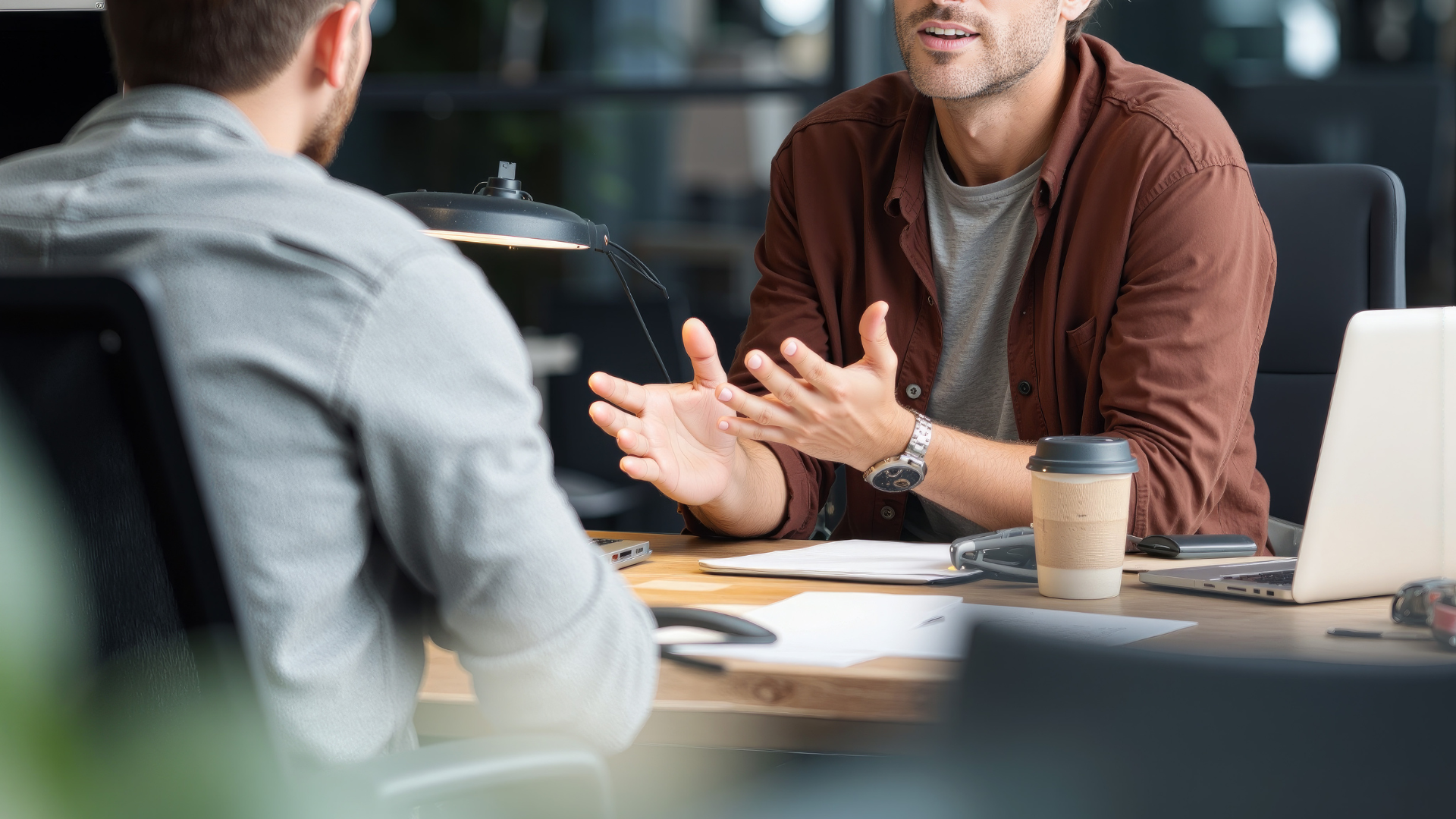 Two men seated at a desk, one gesturing with hands, discussing; papers and coffee cup present.