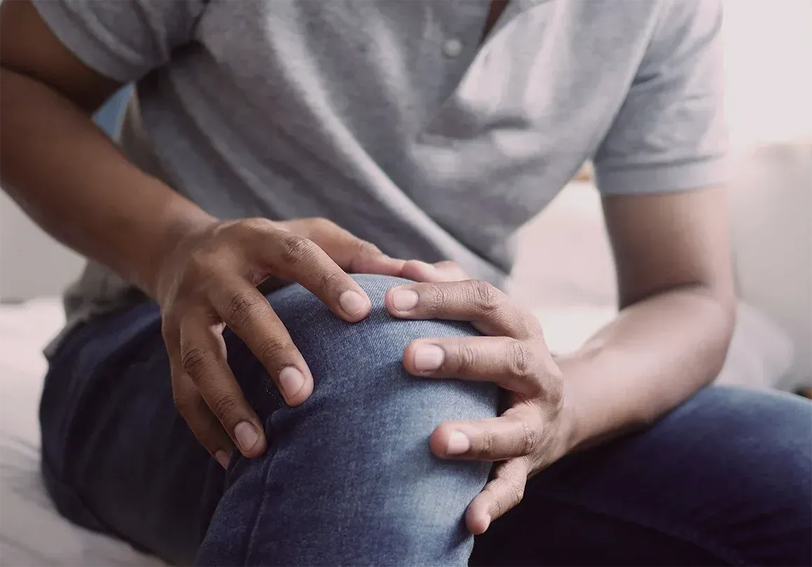 Man holding his knee, suggesting pain or injury. He is wearing jeans and a gray shirt.