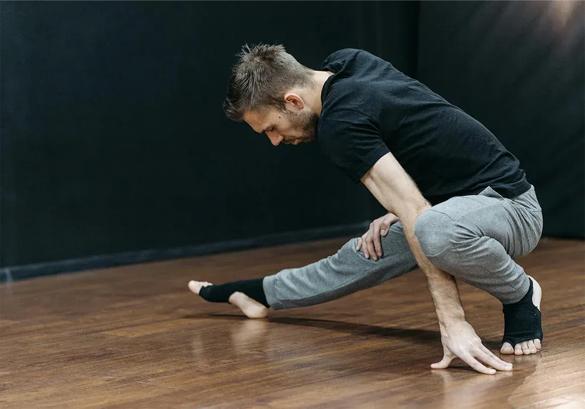Man in athletic wear stretches on wooden floor, hands and foot touching the ground.