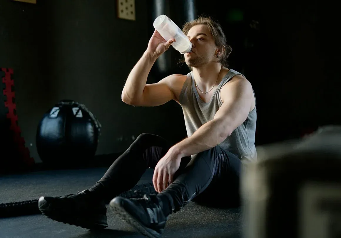 Man in workout clothes, drinking from water bottle, sitting on the floor in a gym.