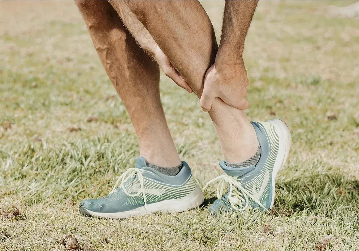 Person holding their ankle in pain outdoors, wearing blue athletic shoes and standing on grass.