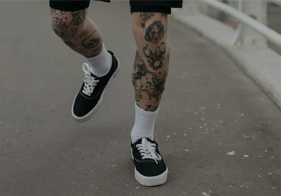 Person with tattooed legs wearing black sneakers, white socks, and shorts, walking on a concrete path.
