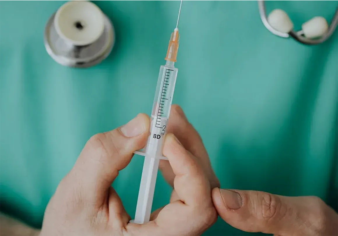 Hands holding a syringe with needle, stethoscope in background. Green scrubs.