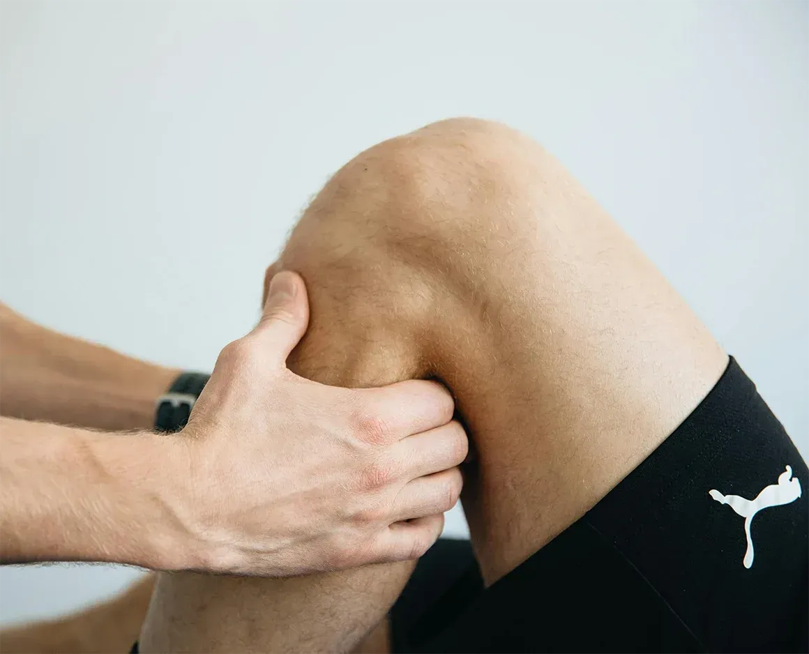Person's knee being examined by another person's hands. White background, black shorts.