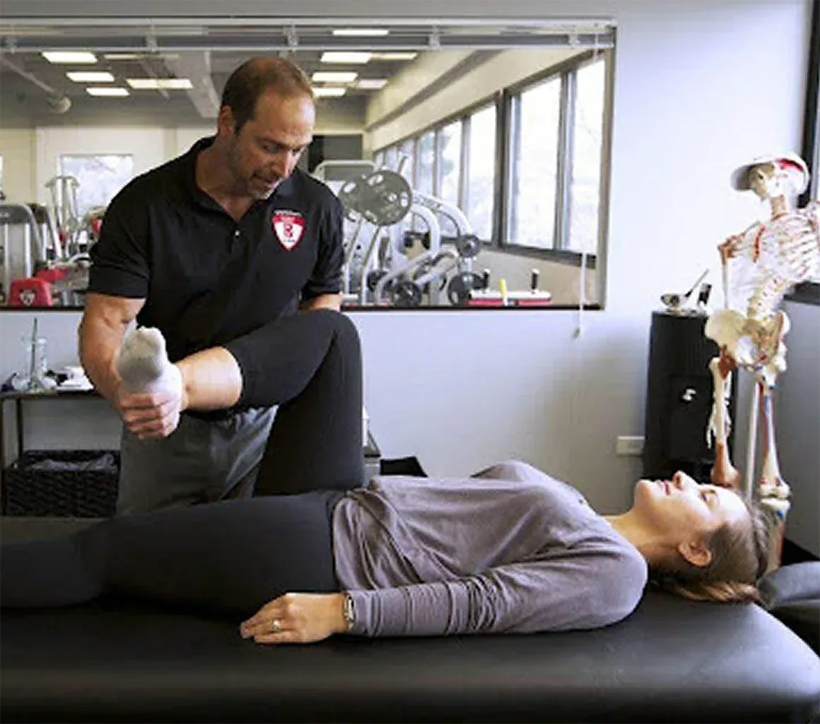 Physical therapist assisting a person on a table with leg stretch; indoor clinic, exercise equipment in background.