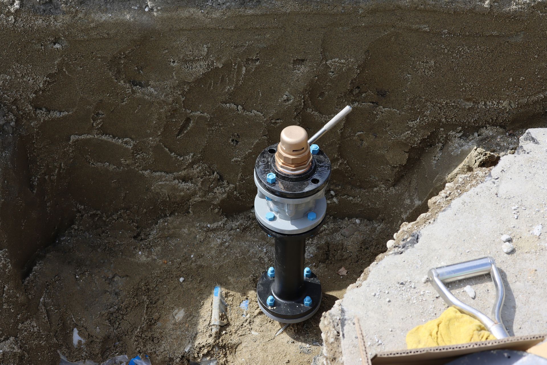 Black pipe with blue bolts, brass top, in a dirt trench, near concrete, with a shovel.