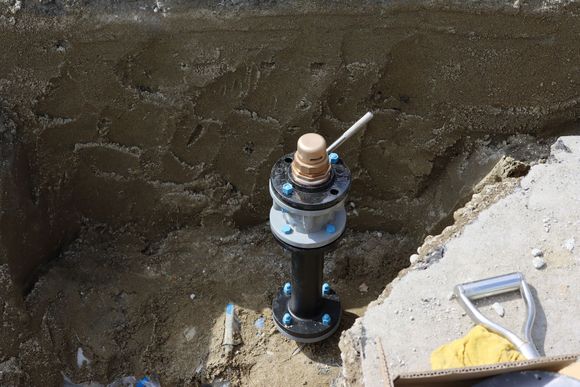 Black pipe with blue bolts, brass top, in a dirt trench, near concrete, with a shovel.