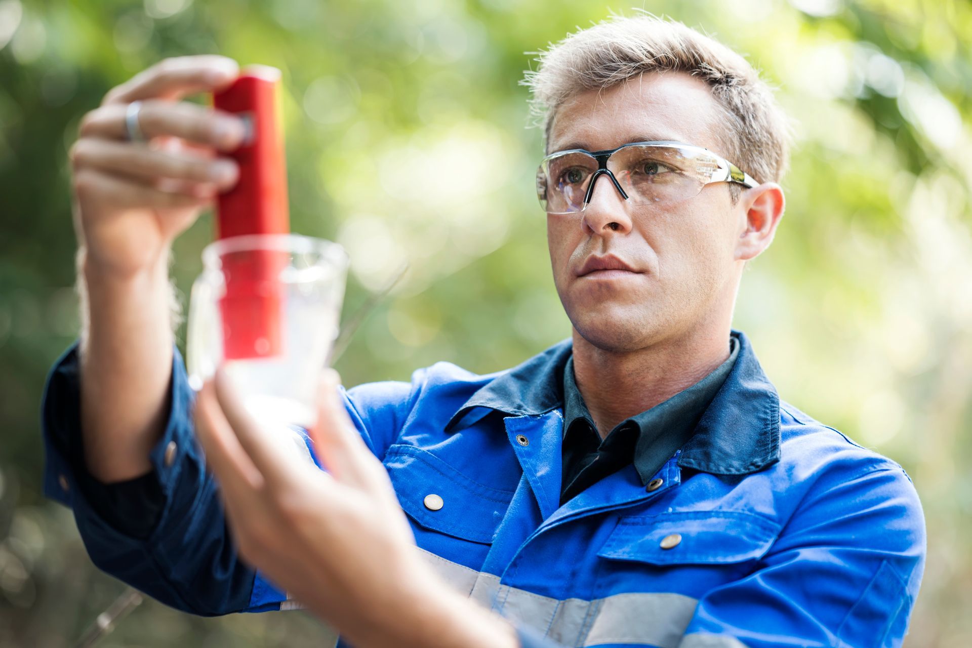 Man in blue overalls tests water sample with a red device outdoors.