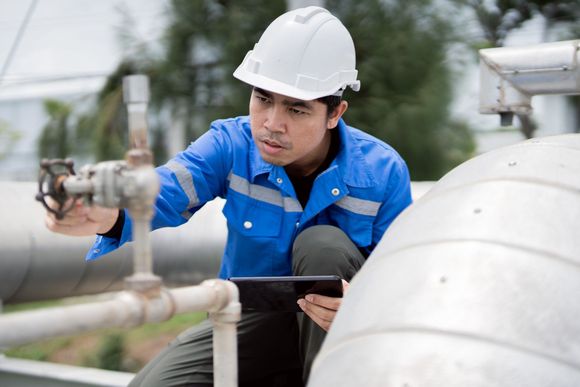 Worker in blue coveralls and hard hat inspecting pipes, holding a tablet.