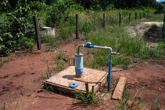 Well pump in a field, with blue pipes and brown earth, surrounded by grass and a fence.