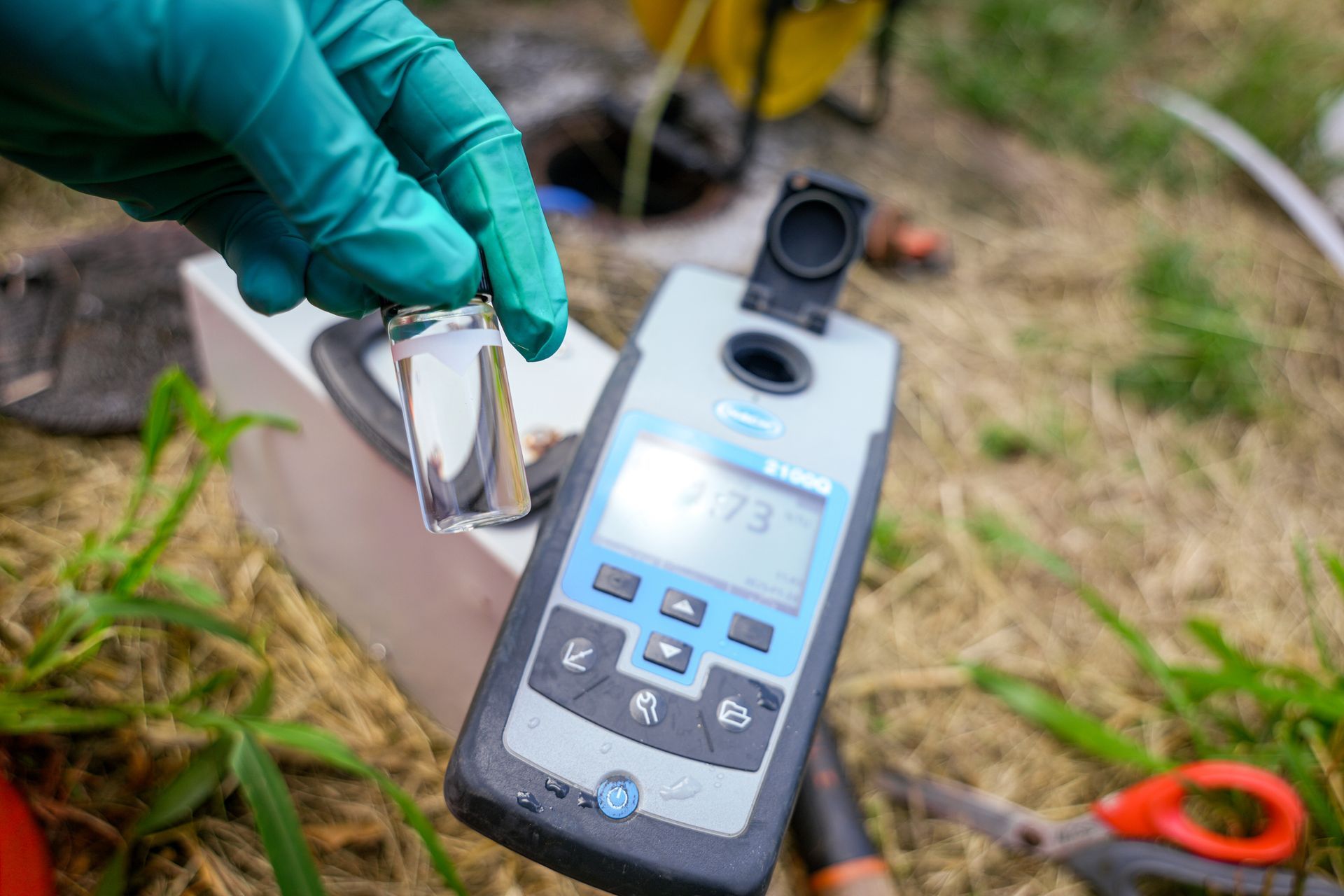 Gloved hand holding a water sample vial near a digital water quality meter outdoors; grass background.