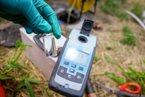 Gloved hand holding a water sample vial near a digital water quality meter outdoors; grass background.