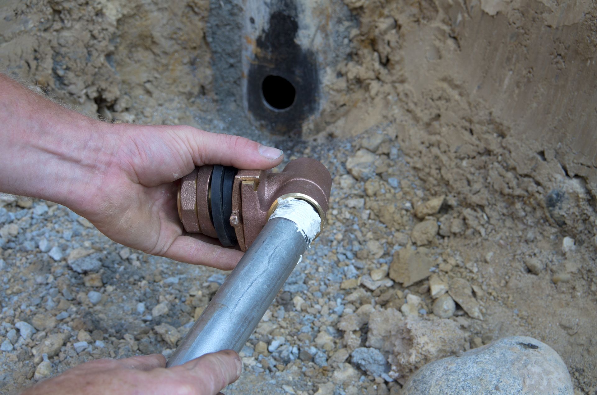 Person connecting a metal pipe to a fitting in a dirt trench.