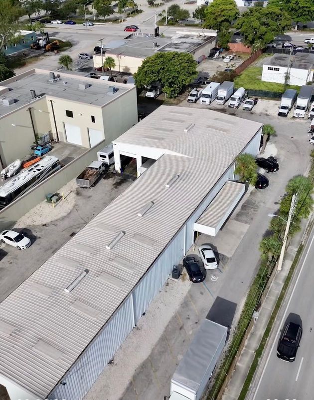 Aerial view of a long industrial building with a corrugated metal roof, cars parked along the street, and some trucks.