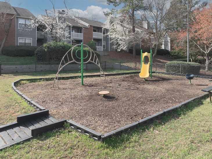 Playground with a slide, and climbing structure, surrounded by mulch and a retaining border, apartments in the background.