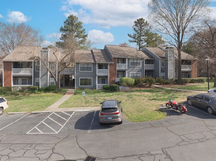 Apartment complex with gray exterior and parking lot.  A motorcycle and cars are parked in front of the building on a sunny day.