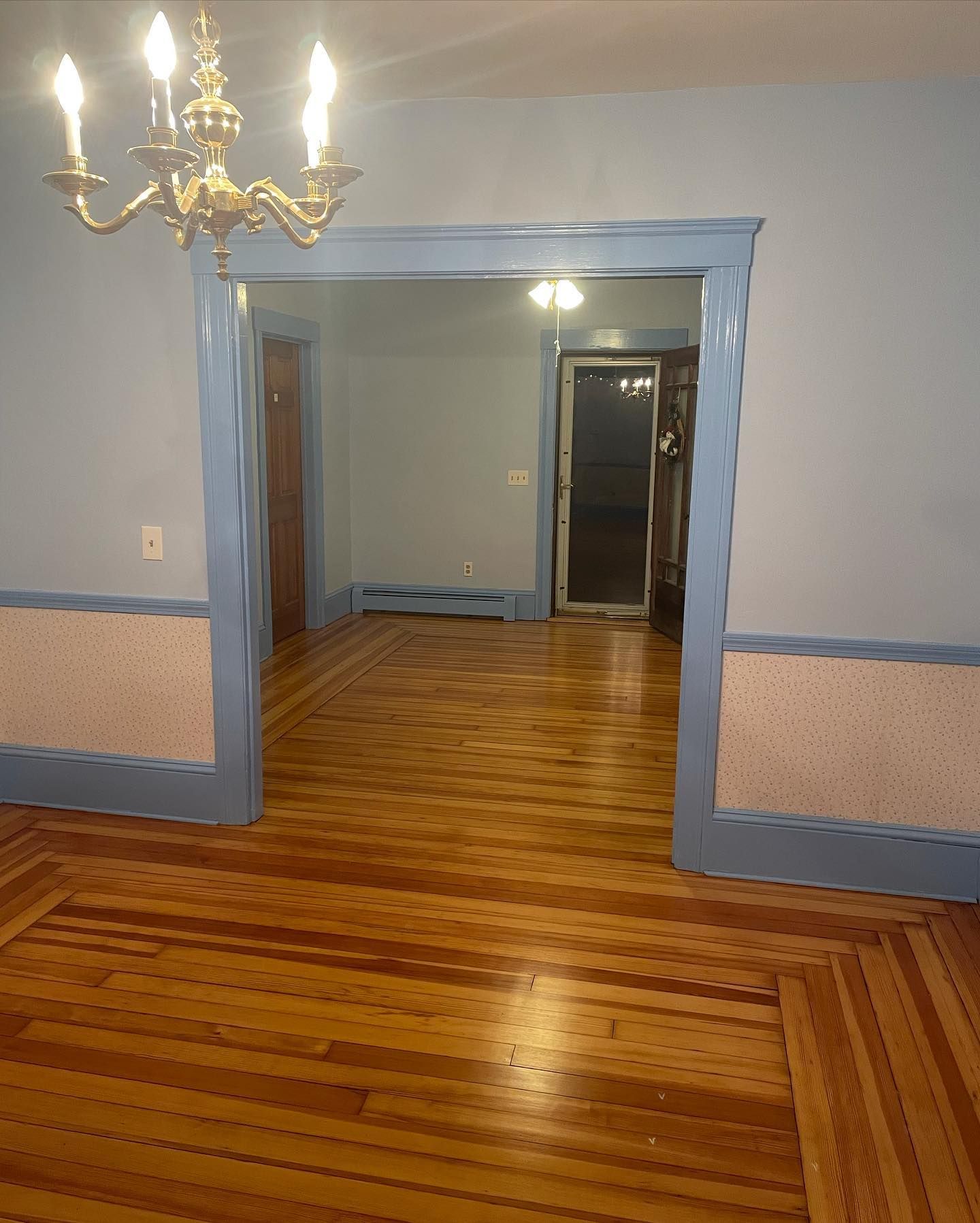 Room with hardwood floors, light blue walls, and a doorway leading to another room. A chandelier hangs above.
