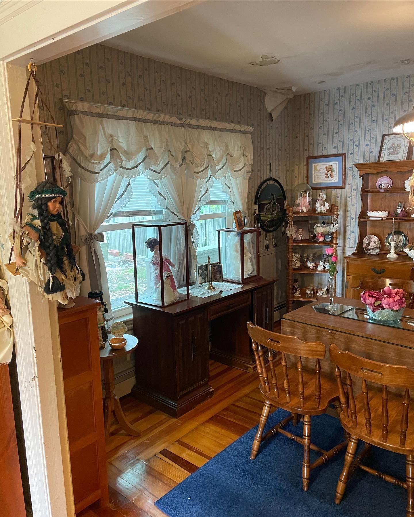 A room with a wooden desk and display cases near a window. Two wooden chairs sit in front of a dining table.