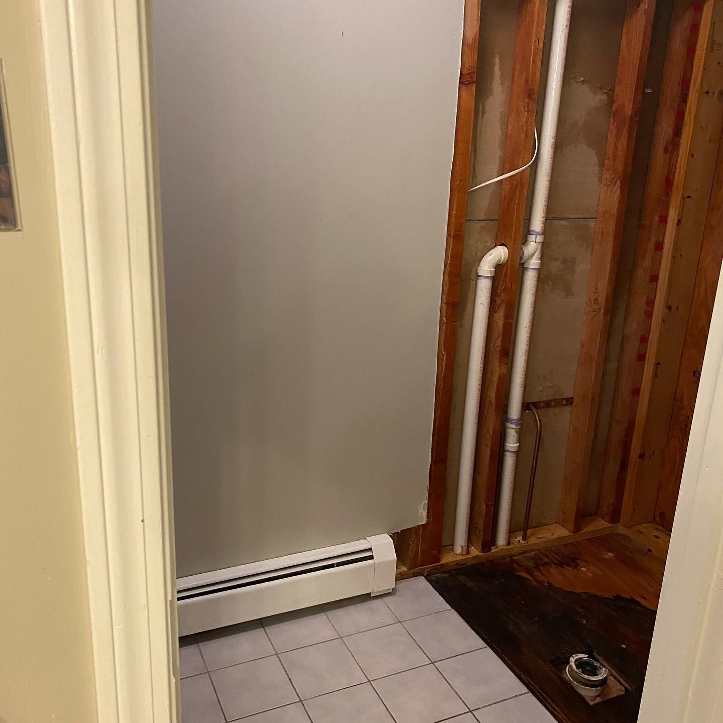Bathroom renovation with exposed pipes, wall studs, and partial flooring. Grey walls, a radiator, and tile visible.