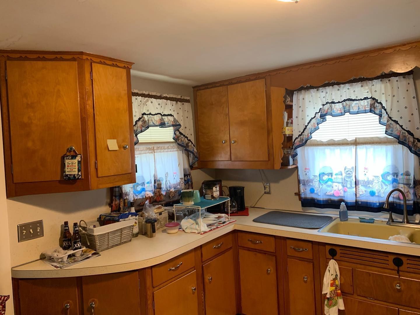 Kitchen with light wood cabinets, white countertops, and small windows with patterned curtains.