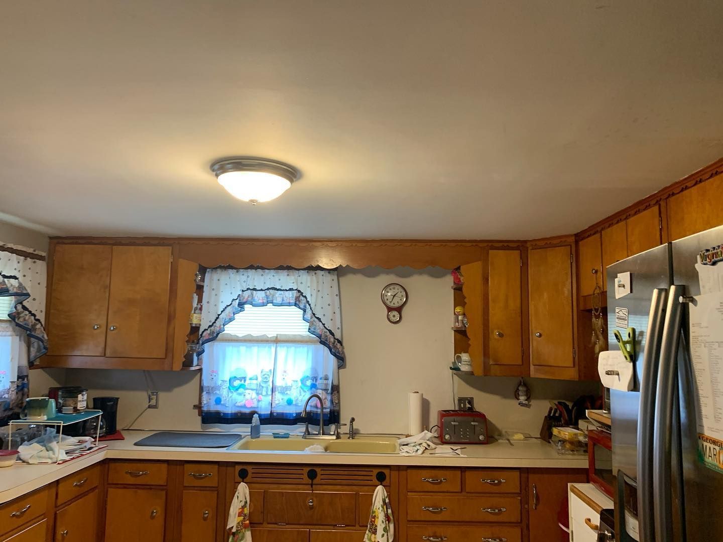 Kitchen with wooden cabinets, a sink, and a window with a curtain. A clock hangs on the wall.