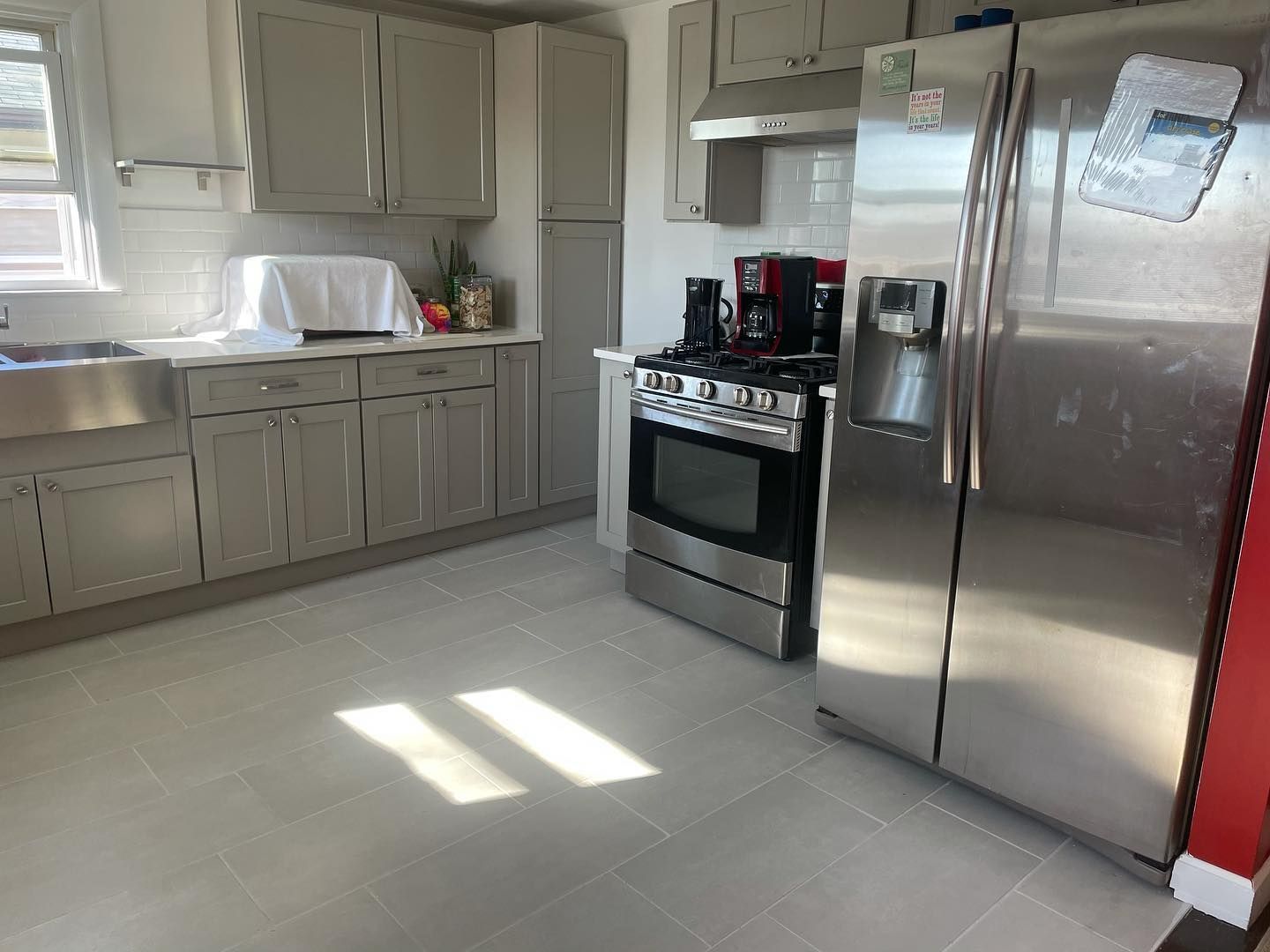 A modern kitchen with gray cabinets, stainless steel appliances, and tile floor. Sunlight streams in.