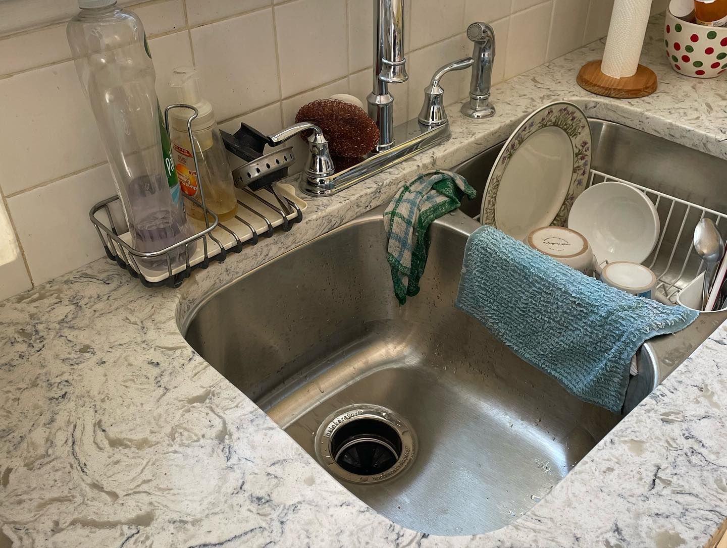 A cluttered kitchen sink with dishes, cleaning supplies, and a dish rack sits on a countertop.