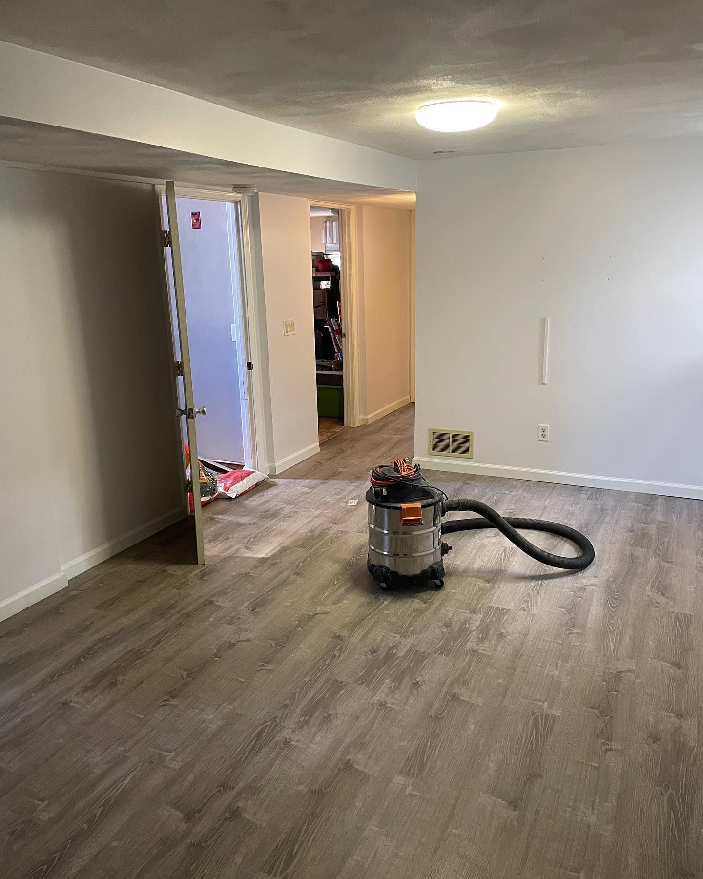 A room with gray wood-look flooring, a vacuum cleaner, and doorways leading to other areas.