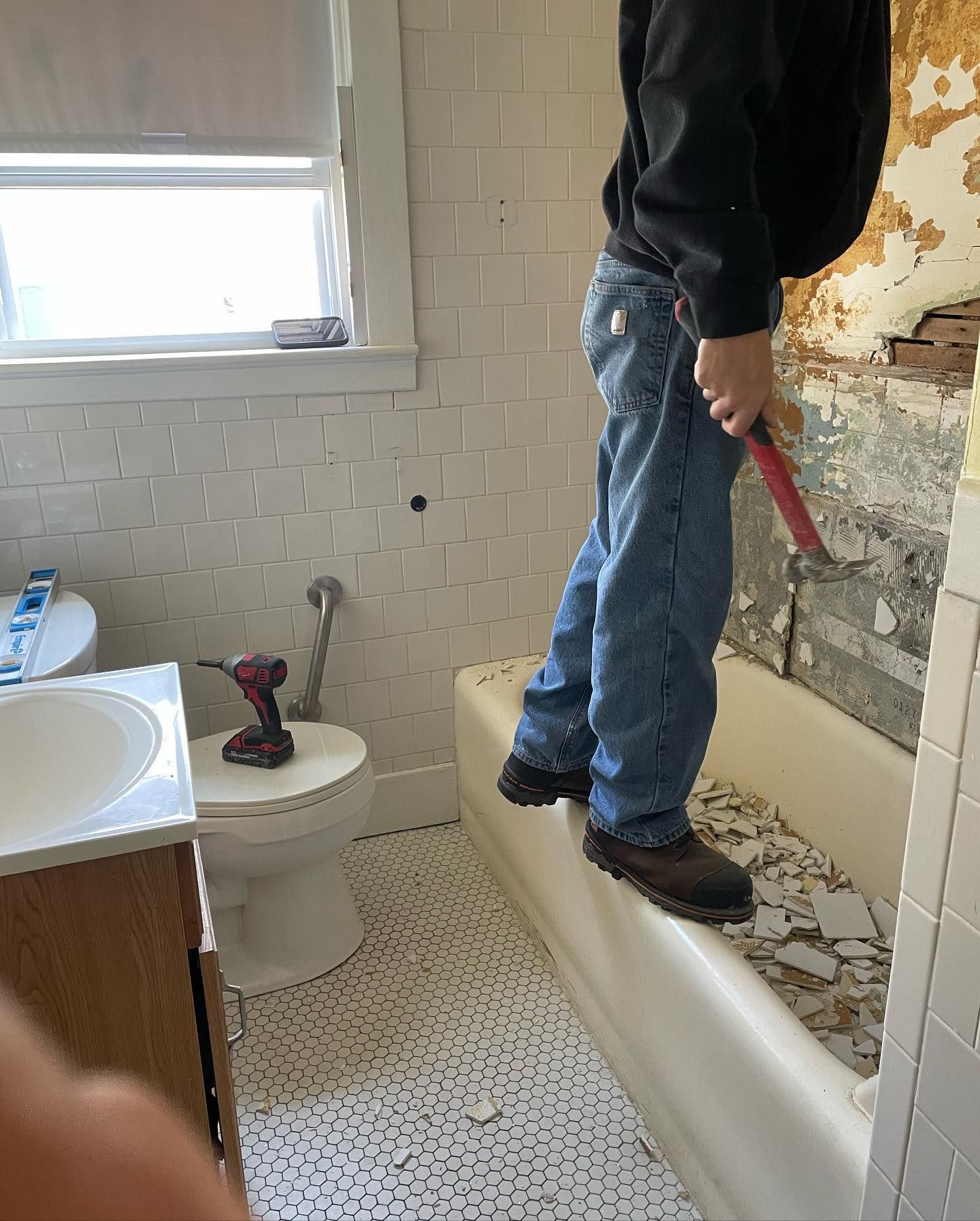 Person removing tile in a bathroom. Standing on bathtub, holding demolition tool. Walls are damaged, toilet