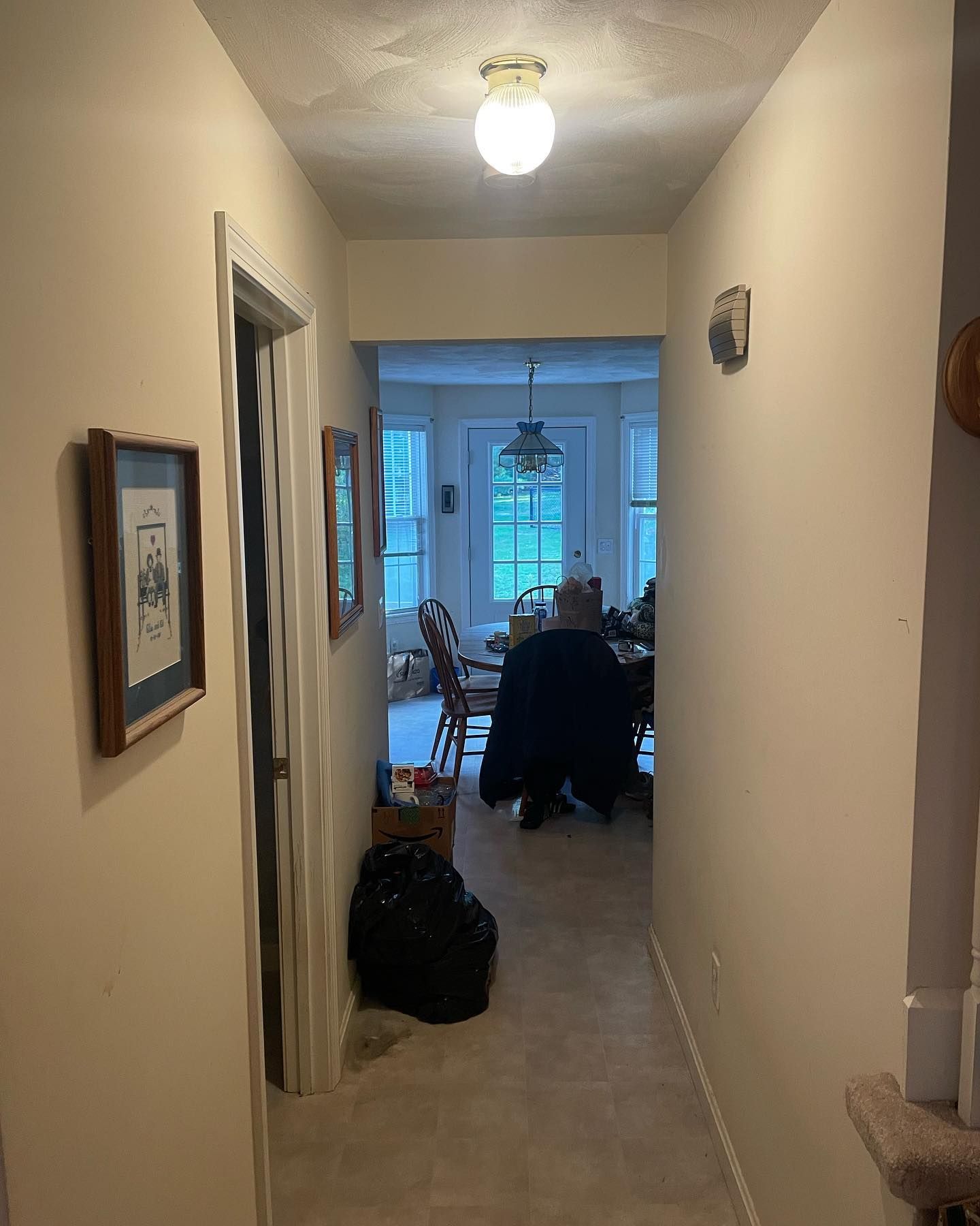 Hallway with light beige walls and a view into a room with windows and a table. A black bag sits on the floor.