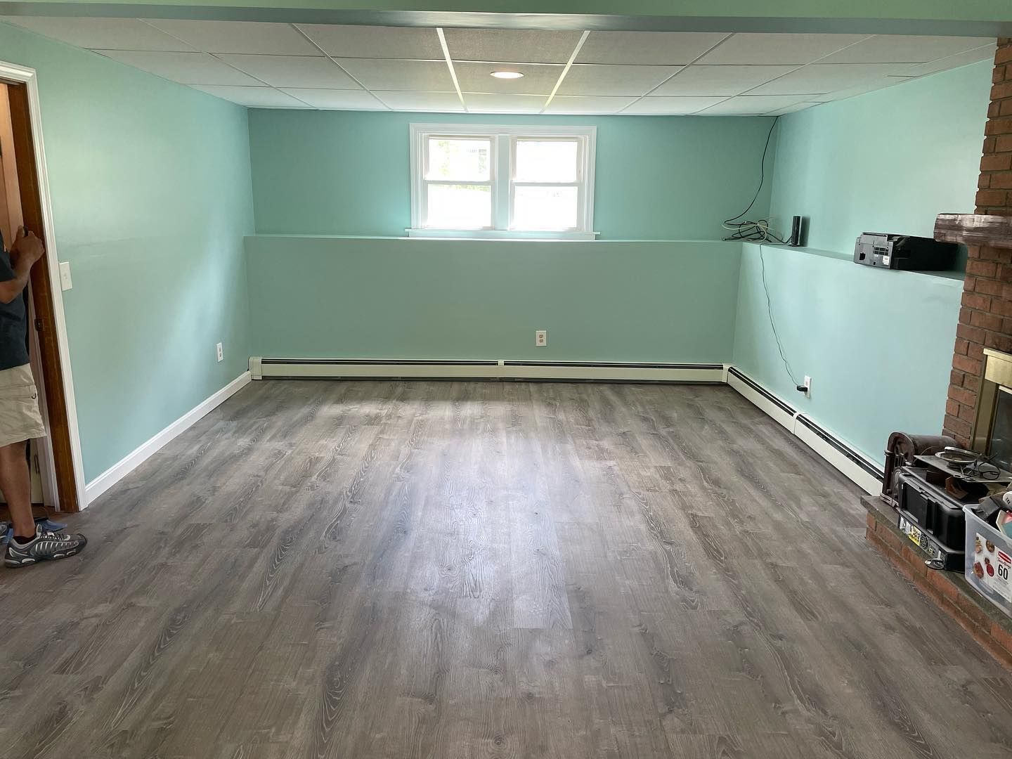 Empty room with teal walls, gray flooring, and a small window. A person stands in the doorway.