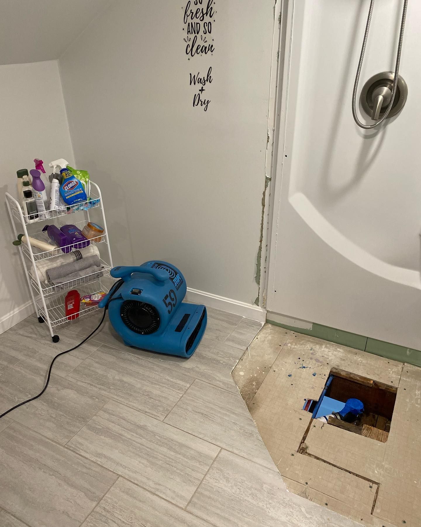Bathroom with exposed floor, drying fan, cleaning supplies on a cart, and shower/tub.