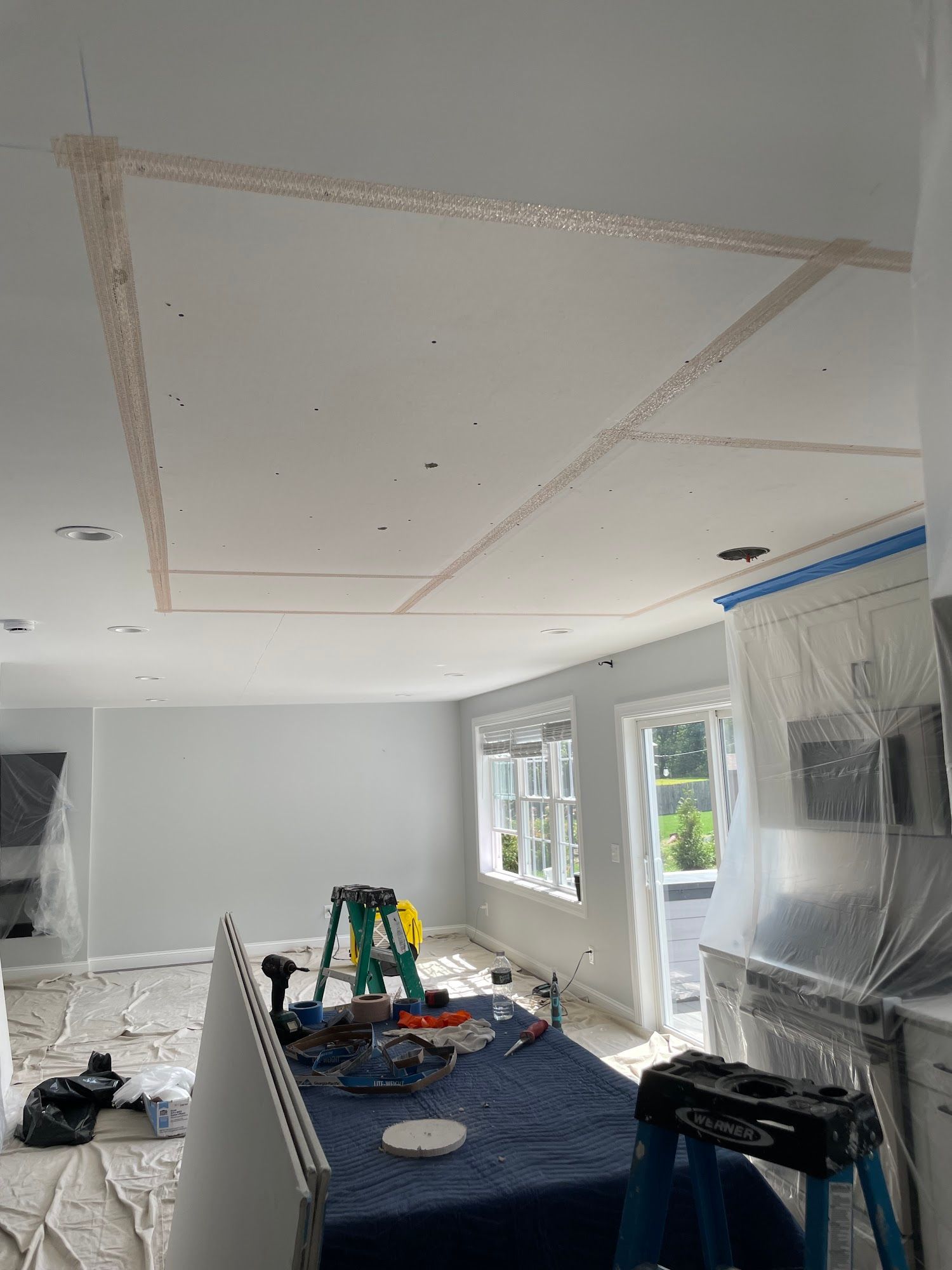 Interior ceiling construction with taped joints, tools, and blue tarp-covered furniture.