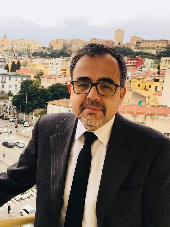 Uomo con occhiali e abito elegante in piedi su un balcone con alle spalle un panorama urbano e edifici in cima a una collina.
