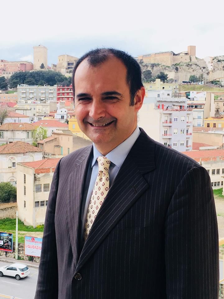 Un uomo sorridente, con indosso un abito gessato scuro e una cravatta a fantasia, si erge di fronte a un paesaggio urbano collinare.